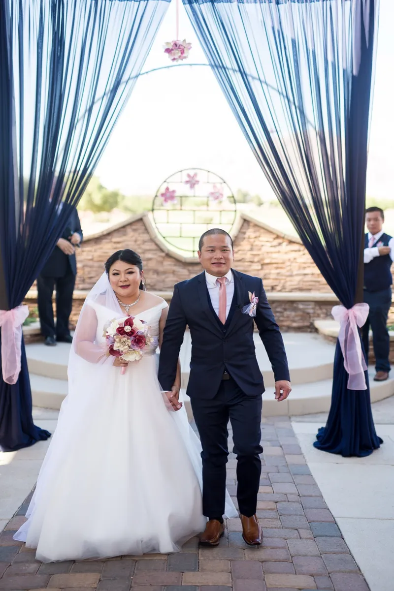 Bride and the older man escorting her walking the paver aisle toward the camera through the navy draped entrance