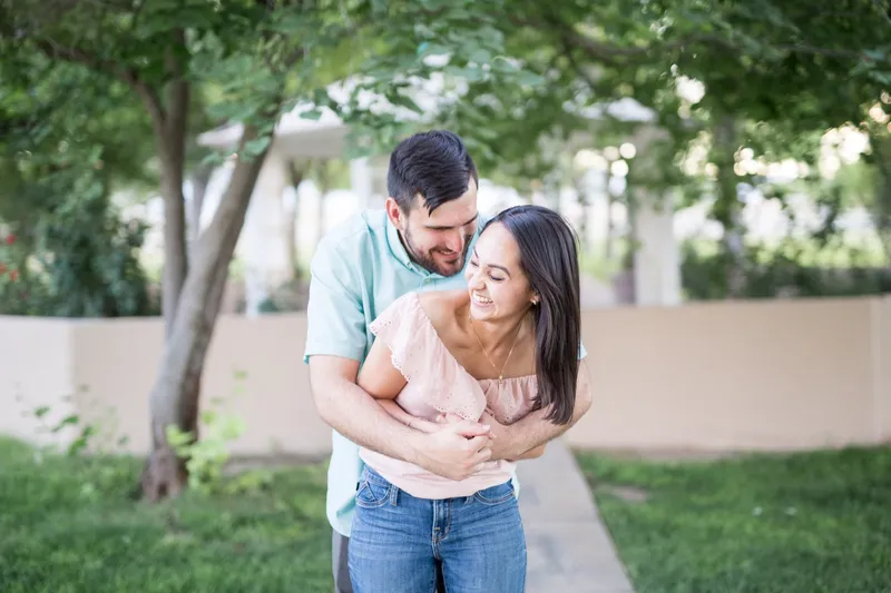 Man hugging his fiancee from behind on the grass while she laughs, with a low wall and trees behind them
