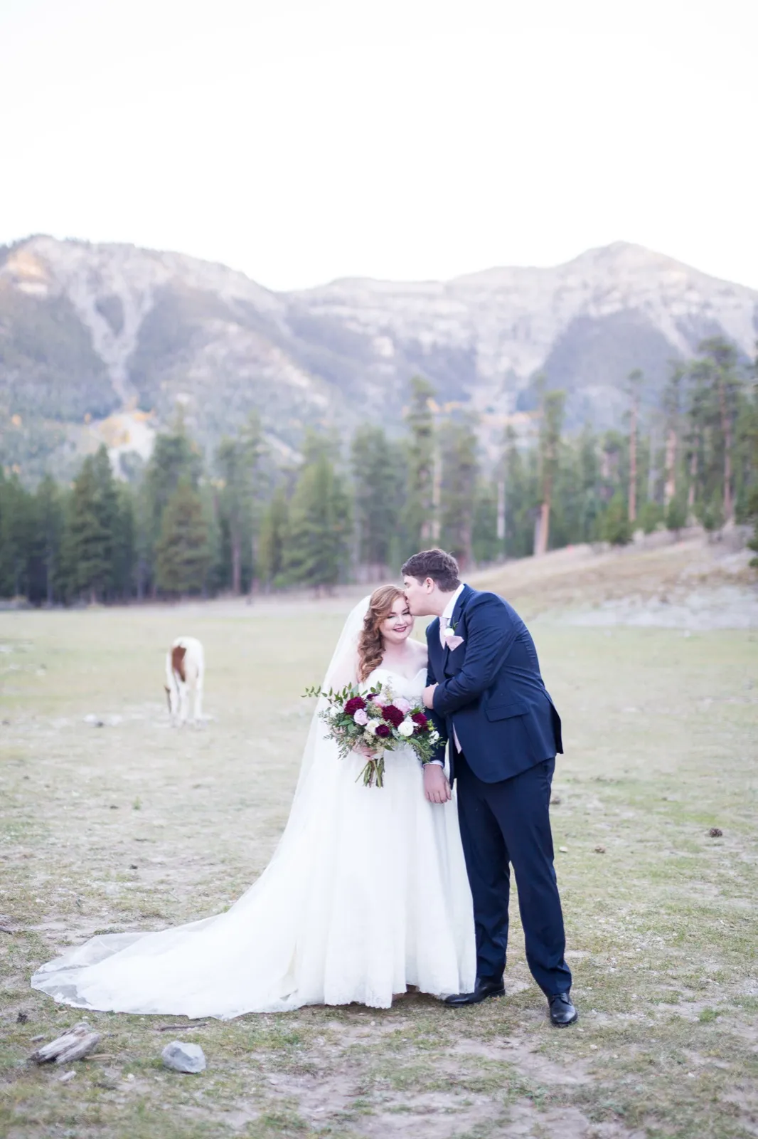 Groom kissing his bride’s forehead in a mountain meadow while a horse grazes behind them under the pines