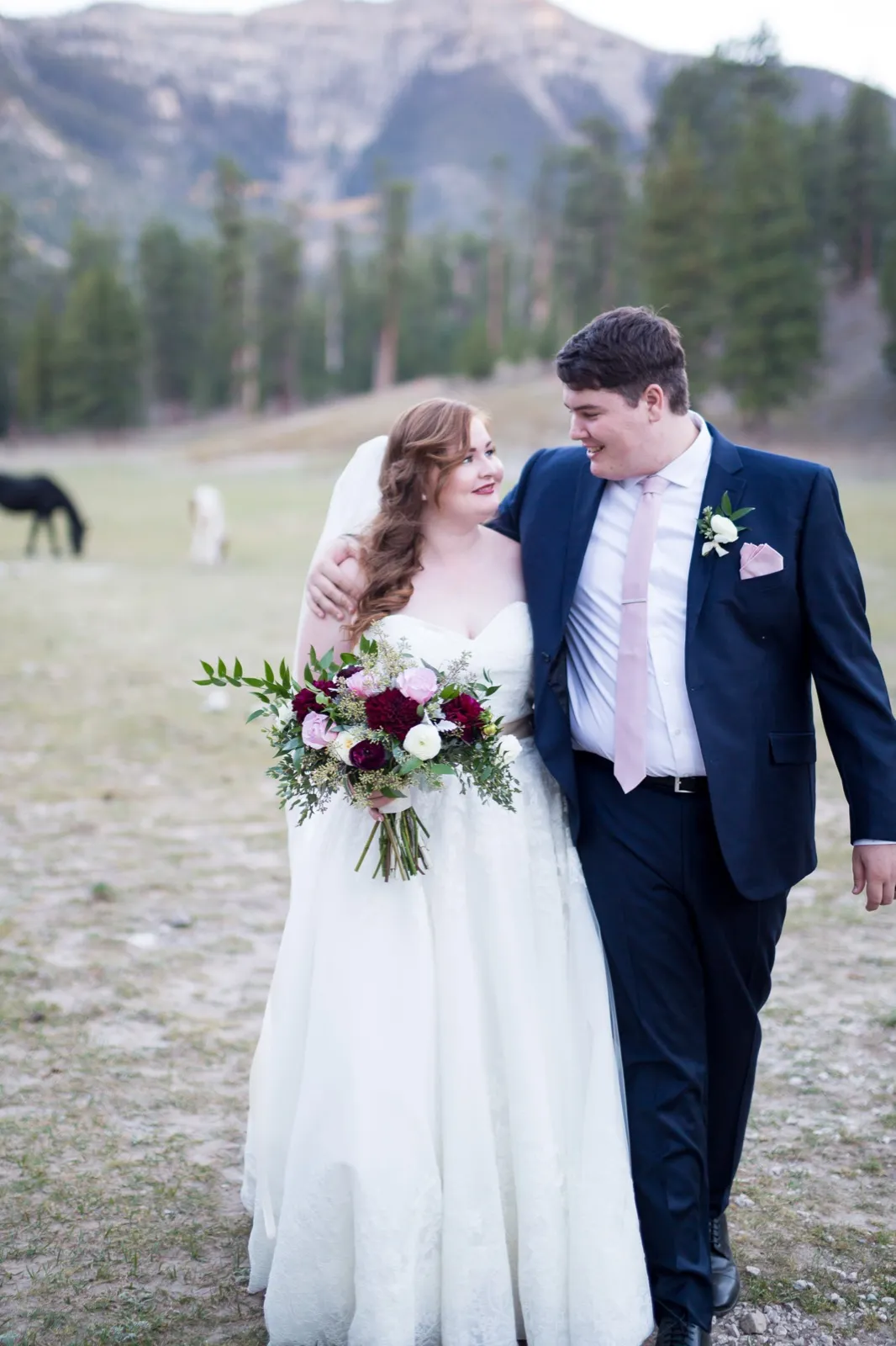 Bride and groom walking arm in arm across a Mount Charleston meadow with horses grazing behind them