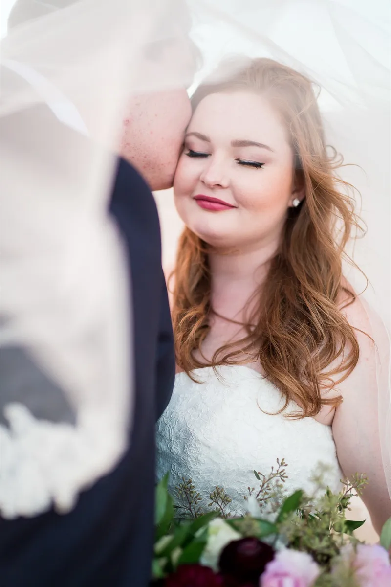 Bride with her eyes closed as her groom kisses her cheek beneath her veil
