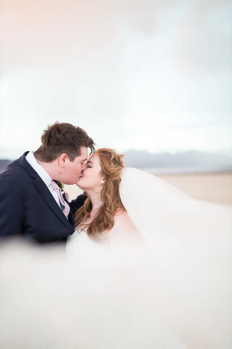 Groom kissing his red haired bride while her long veil blows across the frame on the lake bed