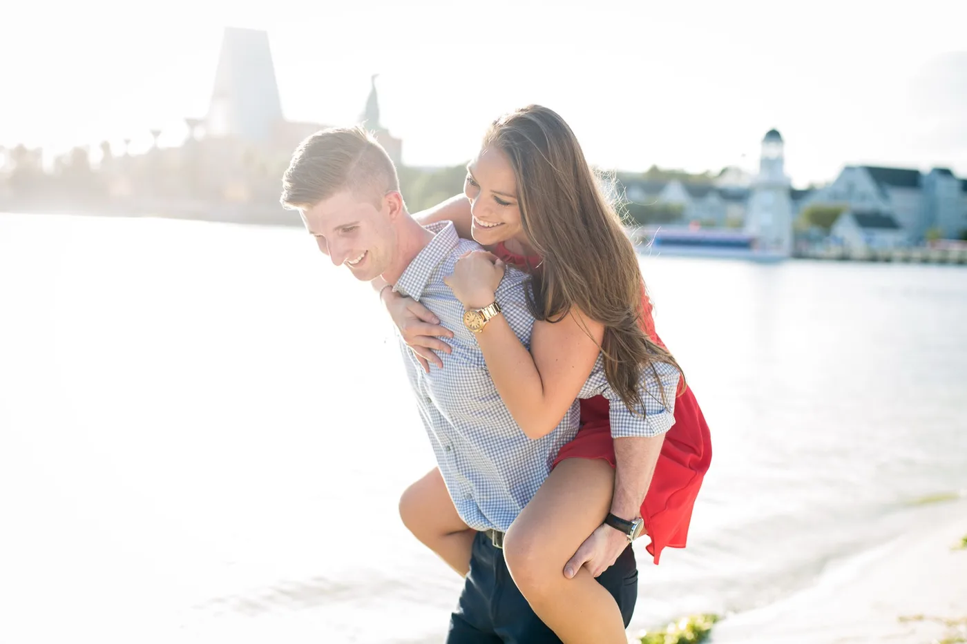 Engaged couple piggybacking along the waterfront at golden hour, both laughing
