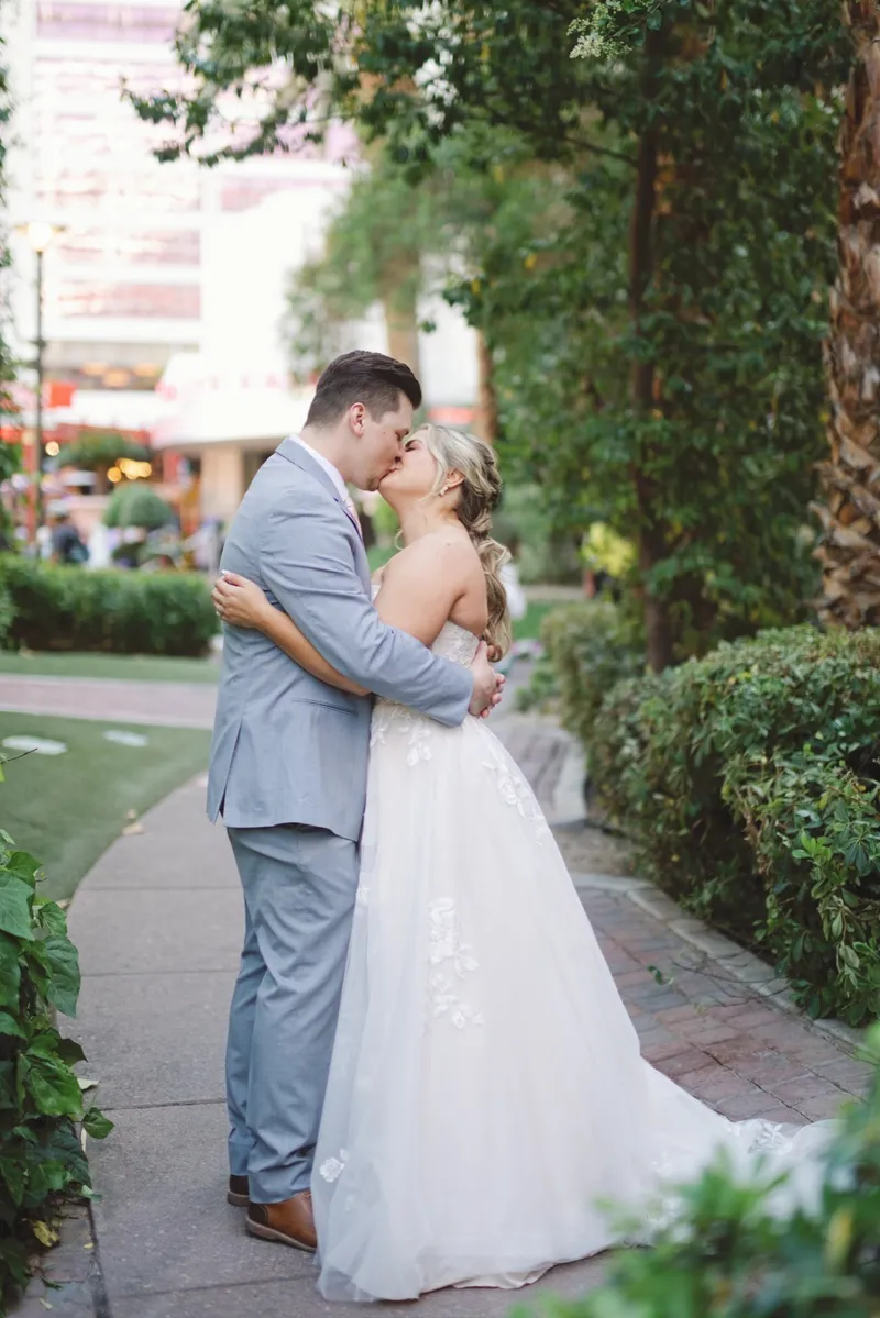 Bride in a lace gown and groom in a light gray suit kissing on the garden path beside a hedge