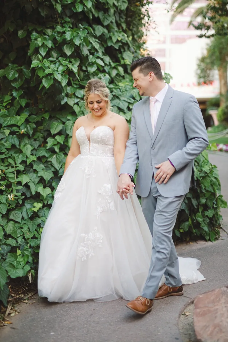 Bride and groom walking hand in hand past a wall of ivy, both of them looking at each other
