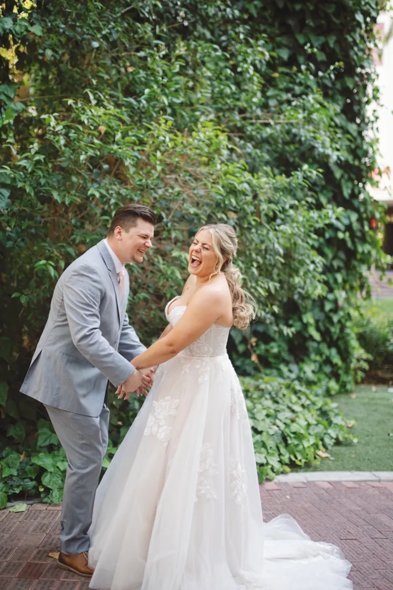 Bride laughing with her head back as the groom holds both her hands in front of the ivy wall