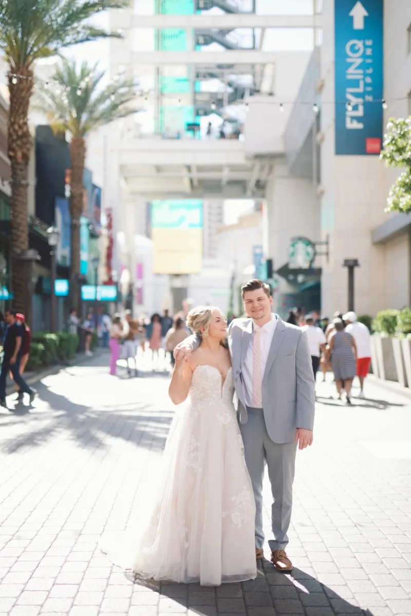 Bride laughing with her hand near her face beside her groom in a grey suit, storefronts and shoppers behind them on the promenade