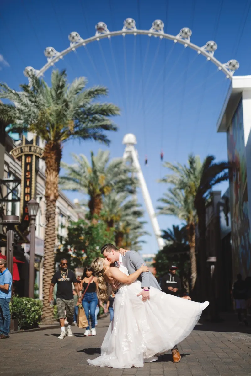 Groom dipping his bride for a kiss on the promenade bricks with the High Roller wheel, palm trees and two zipline riders overhead