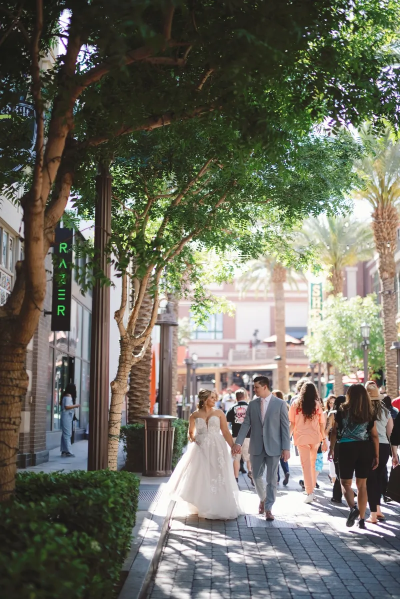 Bride and groom walking away down the tree shaded stretch of the promenade as shoppers pass on both sides