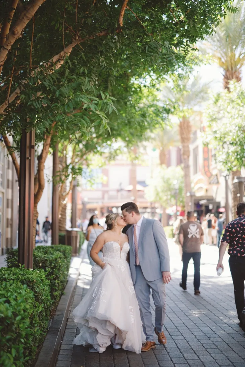 Bride and groom kissing under the promenade trees while people walk past them on both sides