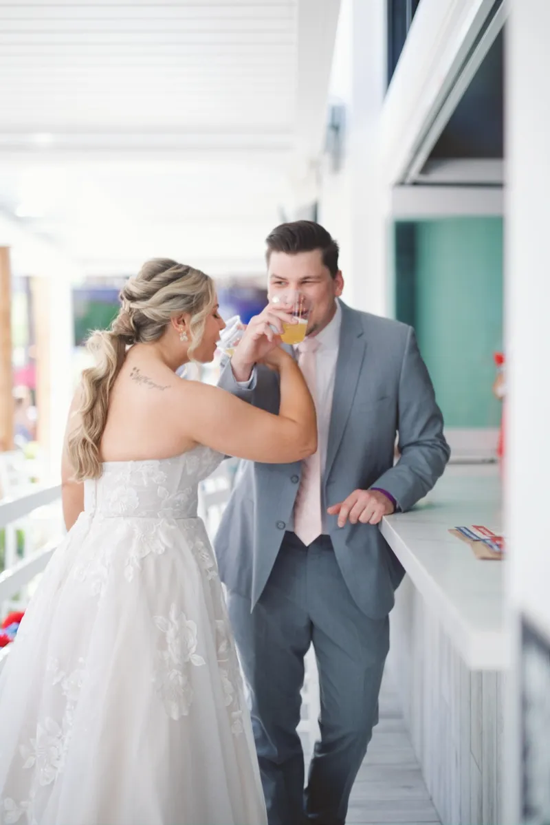 Bride reaching across to drink while her groom sips from his own cup at a white bar counter on the promenade