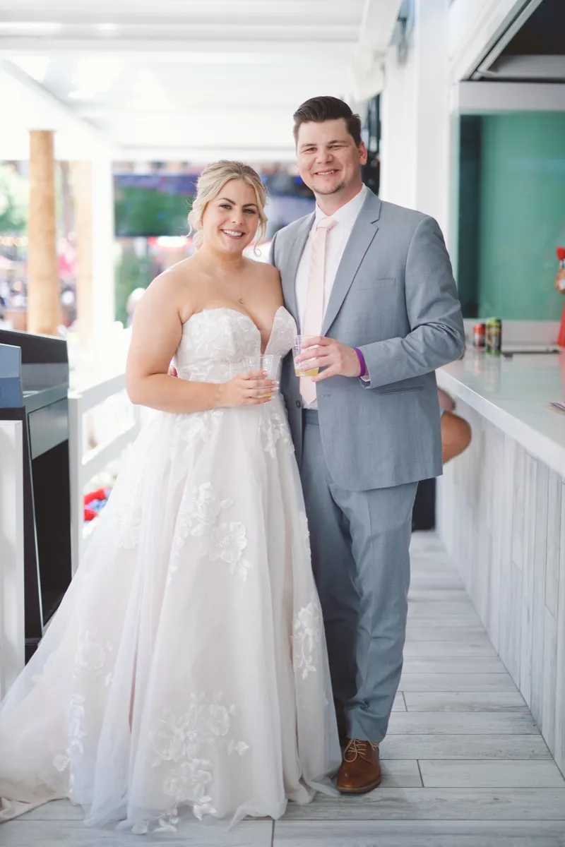 Bride and groom standing at an open air bar counter with drinks in hand, smiling at the camera