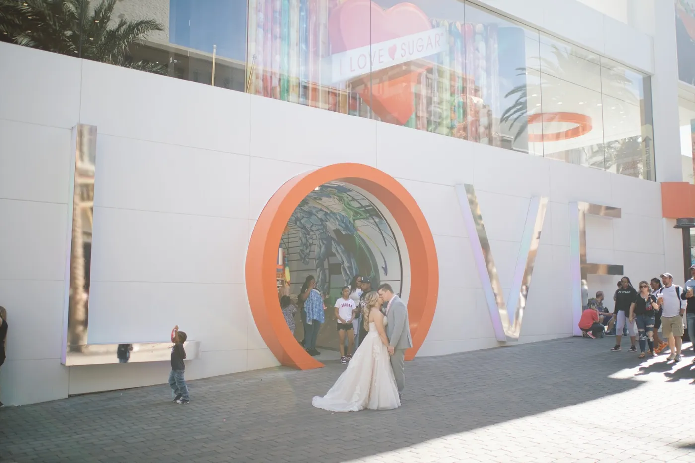 A bride and groom kiss beside giant LOVE letters on a busy promenade while a small boy photographs them