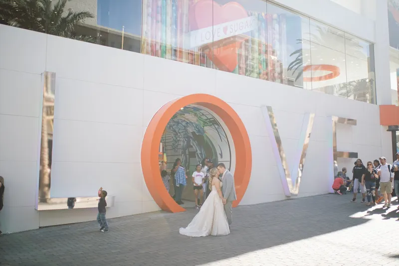 A bride and groom kiss beside giant LOVE letters on a busy promenade while a small boy photographs them