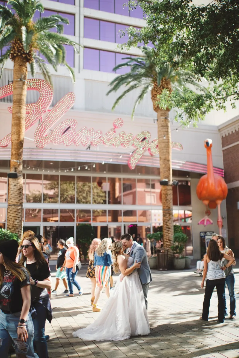 Bride and groom kissing below the Flamingo's pink neon script sign with palm trees, a large flamingo sculpture and a crowd around them