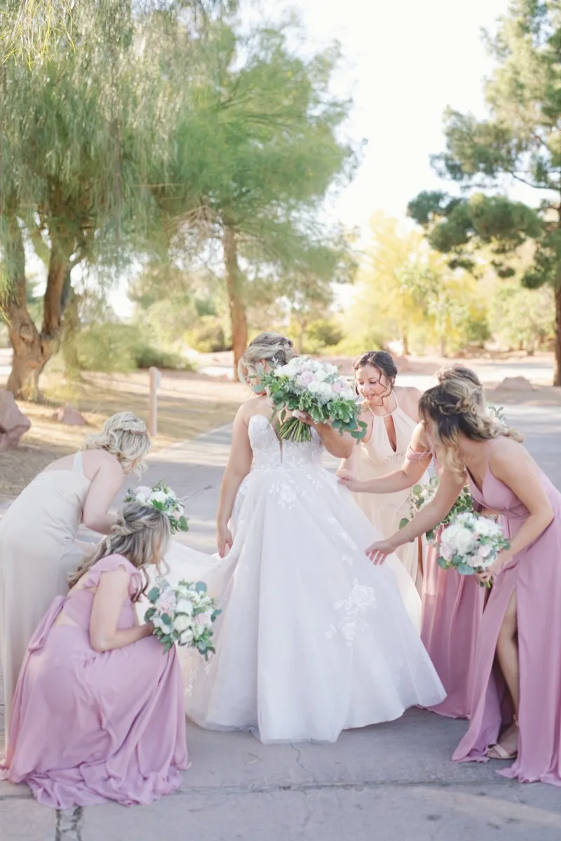 Bridesmaids in dusty pink crouching to fan out the bride's blush gown on the path