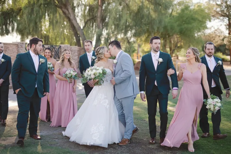 Couple kissing while their wedding party walks alongside them on the grass