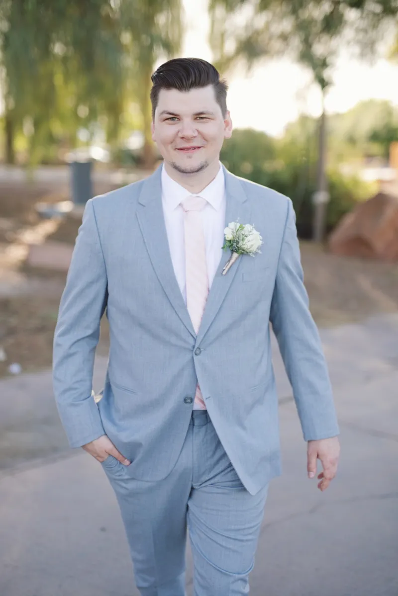 Groom standing alone on the path in a light blue suit with a blush tie, hands in his pockets