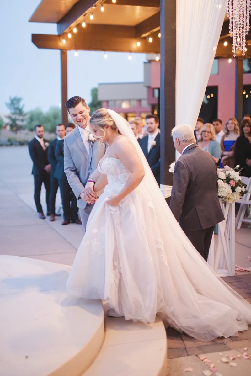Bride in a blush ballgown waiting at the edge of the aisle with her father while the groom and his groomsmen stand under the string lights