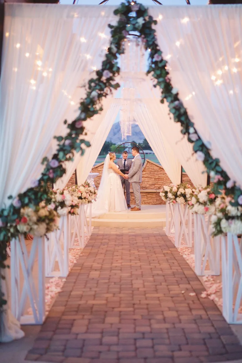 Brick aisle running between two draped pillars wrapped in greenery and flowers with warm string lights overhead, the ceremony underway at the far end