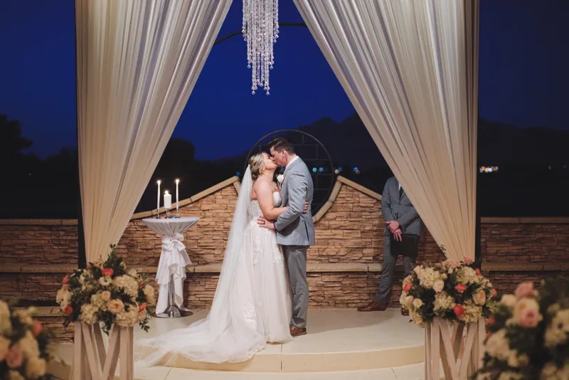 Couple kissing under the draped ceremony structure at dusk, a chandelier hanging above them and lanterns down the aisle