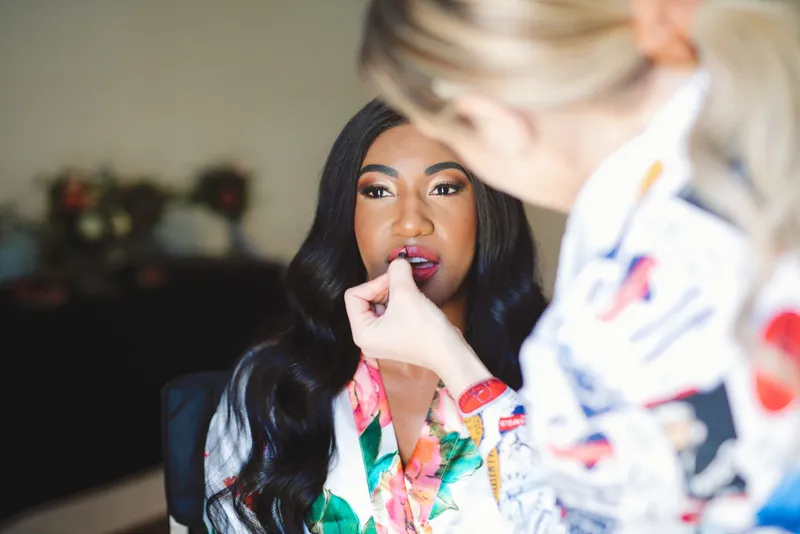 Makeup artist leaning in to paint the bride's lipstick while she sits in a floral robe with her hair loose over one shoulder