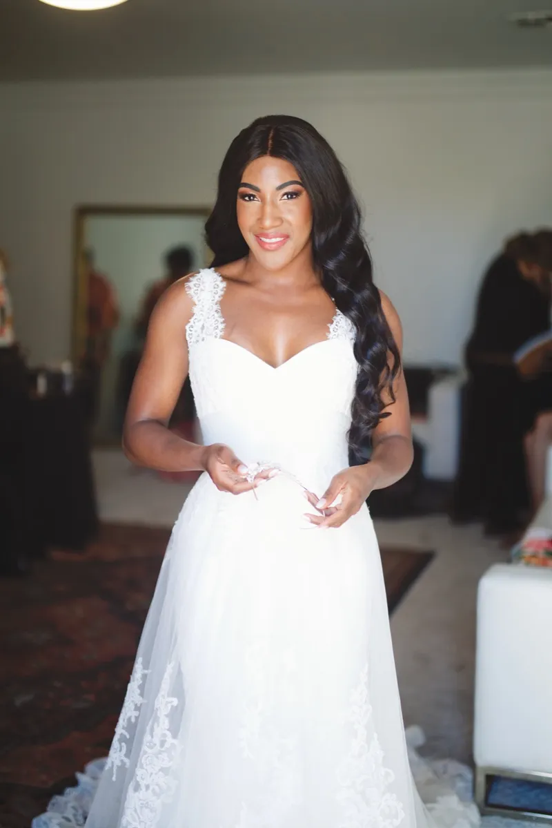 Bride standing in her white lace gown with her hands at her waist, the getting ready room and a patterned rug behind her