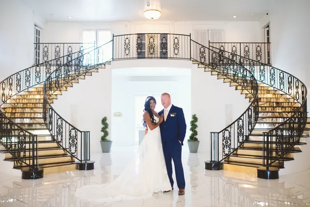 Bride and groom standing together on the polished marble floor of the entry hall, a pair of curved black iron staircases sweeping up on either side of them to a second floor landing