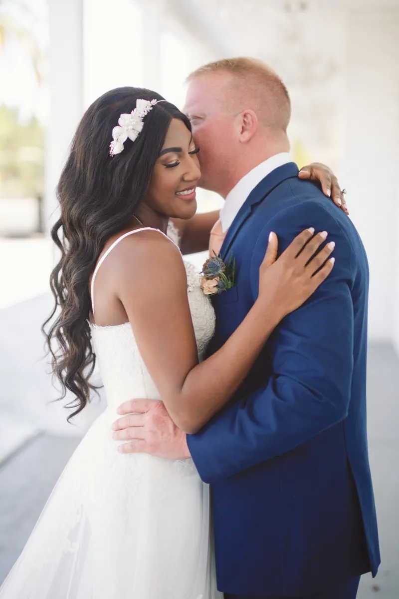 Groom kissing the side of the bride's head while she smiles with her eyes closed, a small flower comb tucked into her hair