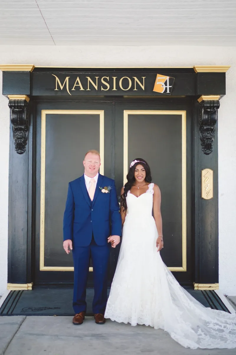 Bride and groom standing in front of the venue's tall black doors, the name Mansion 54 spelled out in gold letters above them