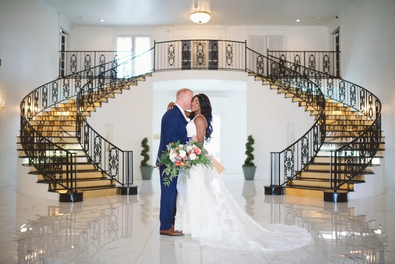 Groom kissing the bride in the entry hall with her bouquet held between them and the iron railings of the staircases filling the frame behind