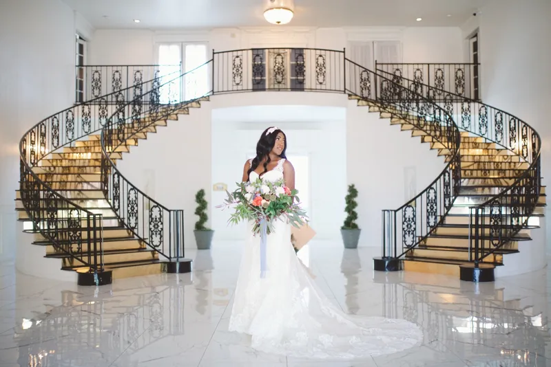 Bride alone in the middle of the entry hall holding her bouquet low, both curved staircases behind her and the marble floor reflecting the light