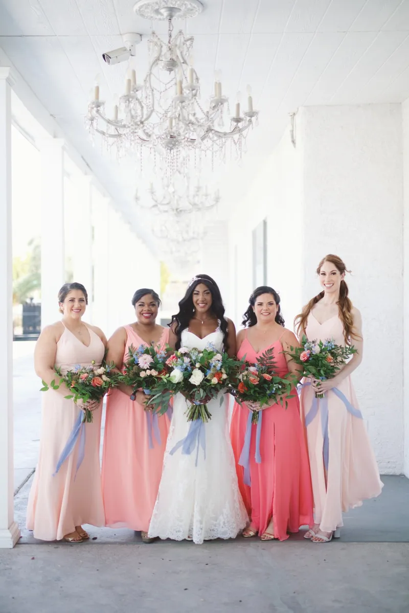 Bride with four bridesmaids in coral and blush gowns, all of them holding bouquets trailing pale blue ribbon, under the chandeliers of the covered walkway