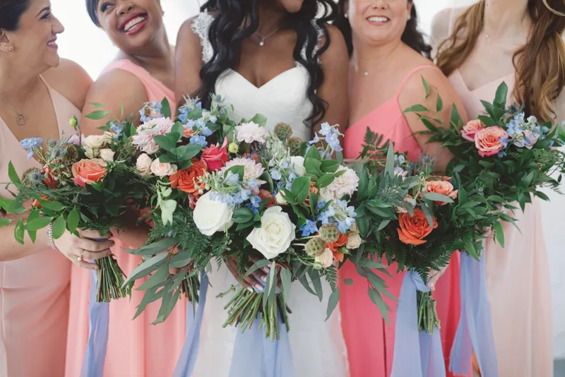 Row of bouquets held together at waist height, coral and cream flowers with greenery and blue ribbon against the coral and blush dresses