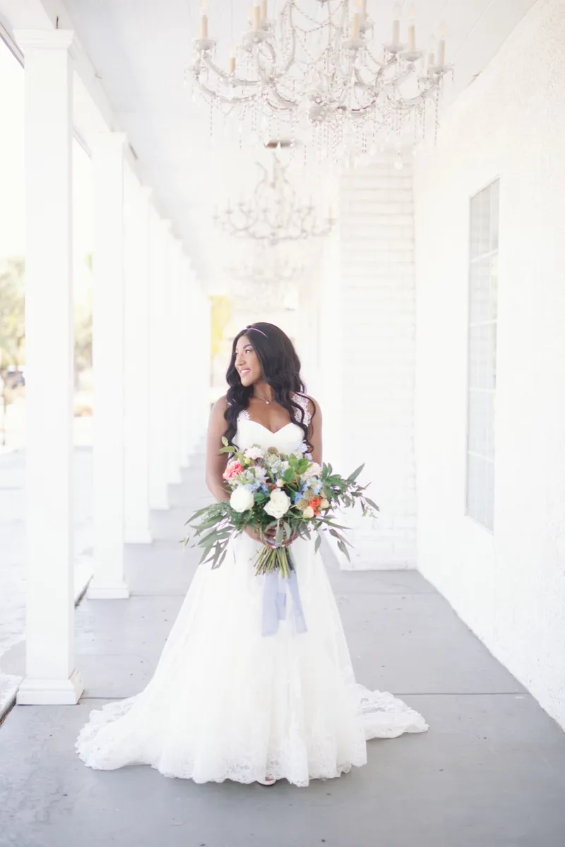 Bride standing in the white covered walkway with her bouquet, a line of chandeliers hanging down the ceiling above her