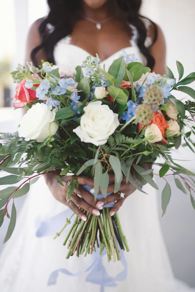 Close view of the bride's hands around a bouquet of cream roses, coral blooms and blue delphinium tied with pale blue ribbon