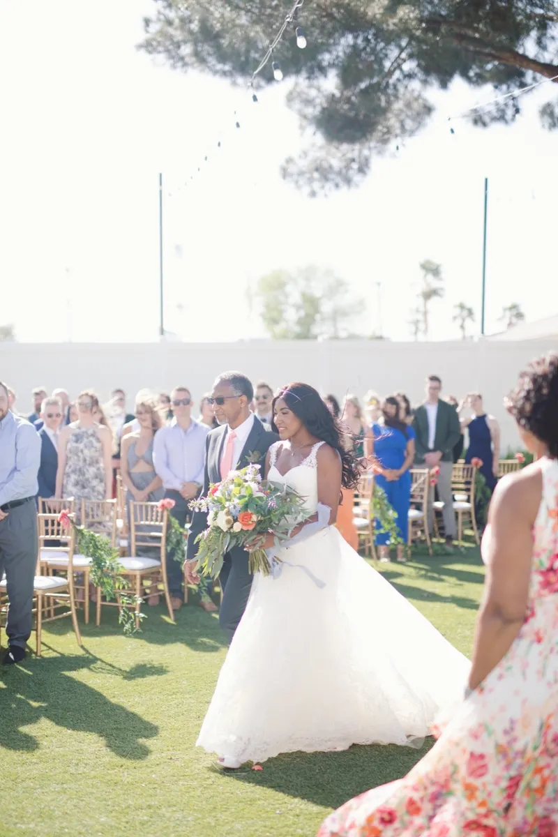 Bride walking up the grass aisle on an older man's arm while guests stand at gold chairs trimmed with greenery