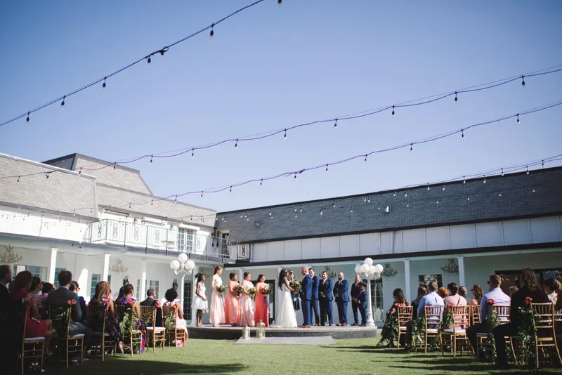 Wide view of the ceremony in the courtyard, guests seated in gold chairs on the lawn and the wedding party on a round platform, with the two story house and strings of cafe lights above them