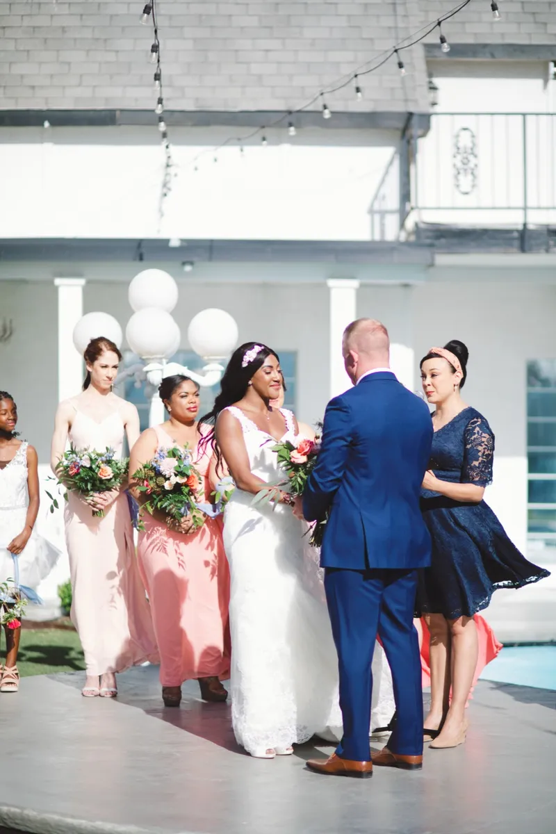 Bride and groom facing each other on the ceremony platform, bridesmaids in blush and coral close beside them and the officiant standing to the groom's left