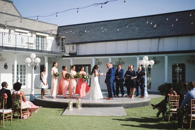 The couple reading vows on the round platform with the swimming pool just behind them, bridesmaids on one side and groomsmen on the other, white globe lamps at both ends