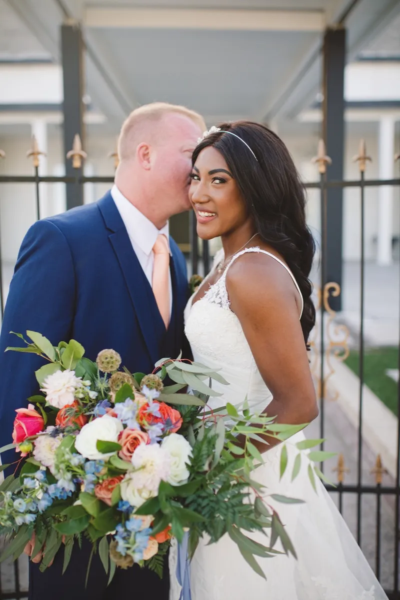 Groom kissing the bride's cheek after the ceremony while she smiles at the camera, her bouquet up at her shoulder and an iron fence behind them