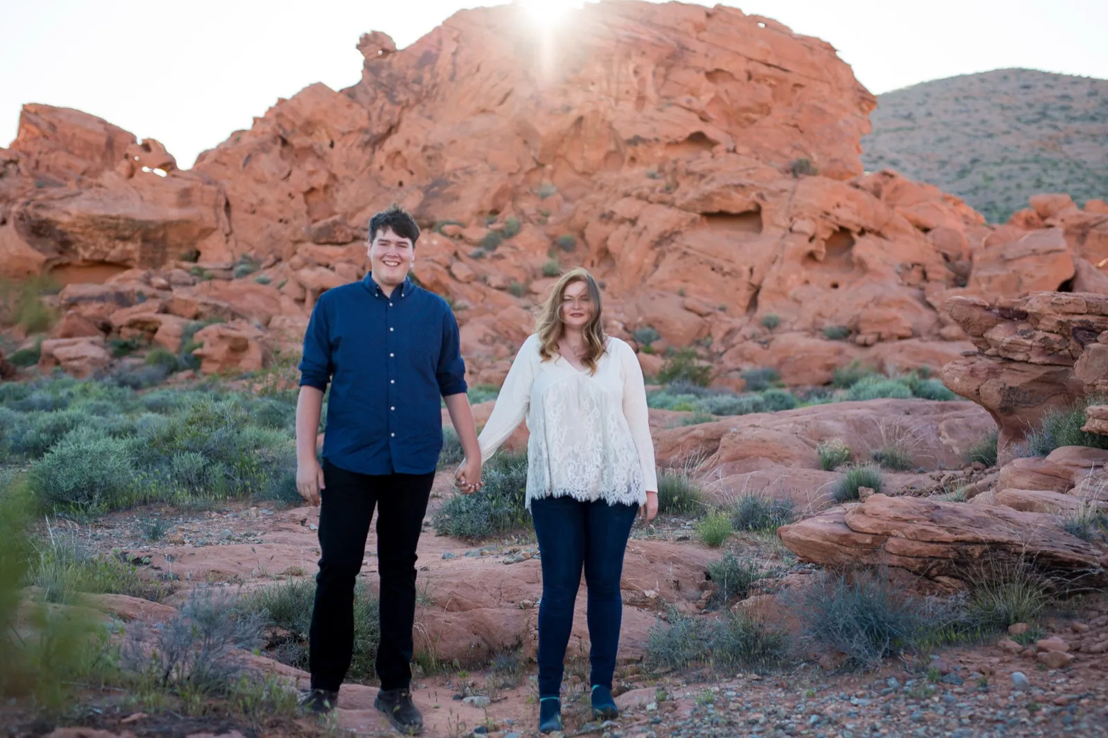 Newly engaged couple hold hands in the sagebrush as the sun crests a red sandstone outcrop at Valley of Fire