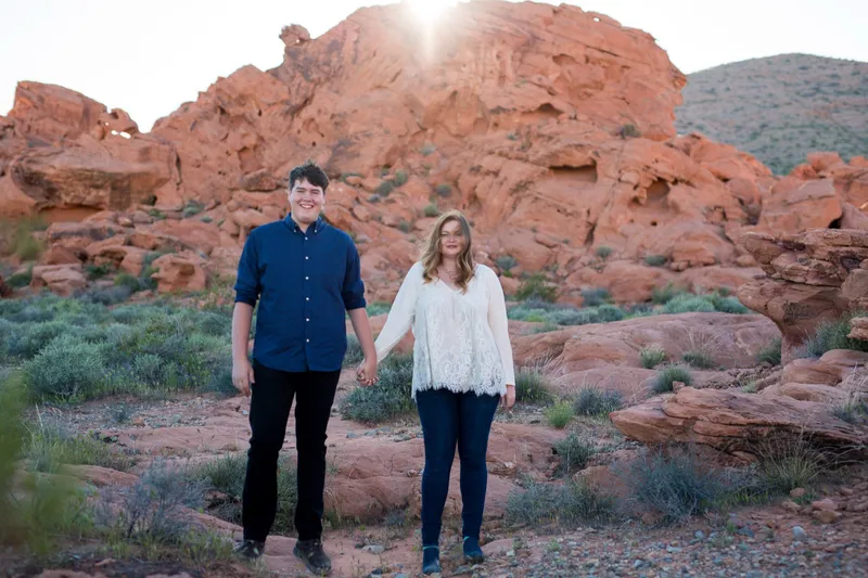 Couple holding hands as they stand together on the desert floor below a red sandstone ridge