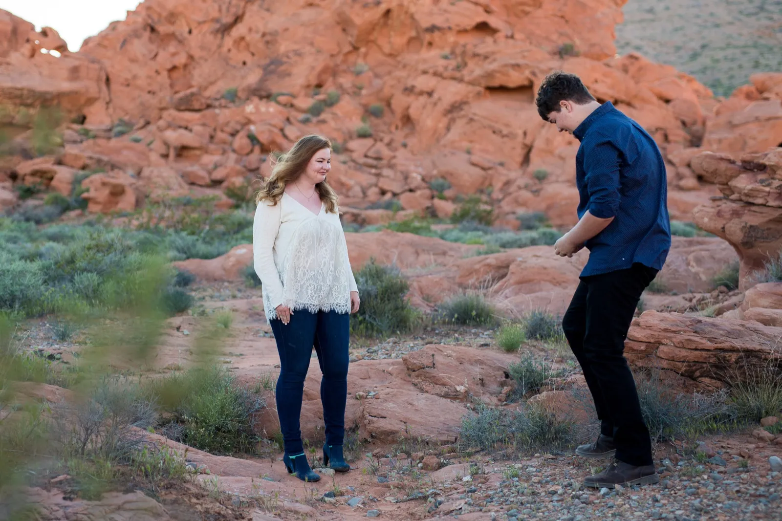 Woman standing on red sandstone looking down and smiling as her partner walks toward her reaching into his pocket