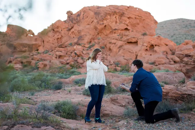 Man down on one knee in front of a red sandstone wall while his partner brings both hands to her face