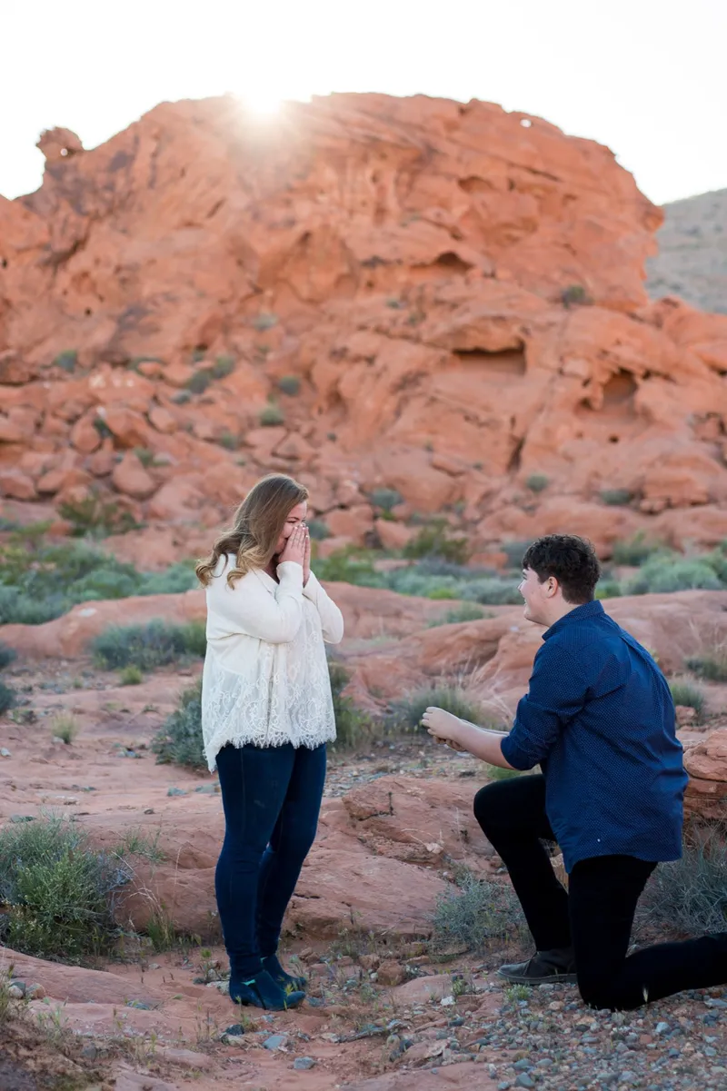 She covers her face with both hands as he kneels in front of her on the red rock