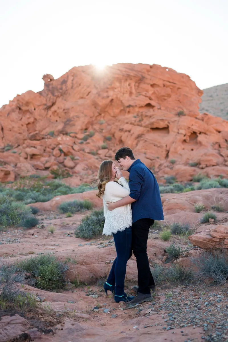 She holds her hands to her face while he wraps both arms around her, sun cresting the rock above