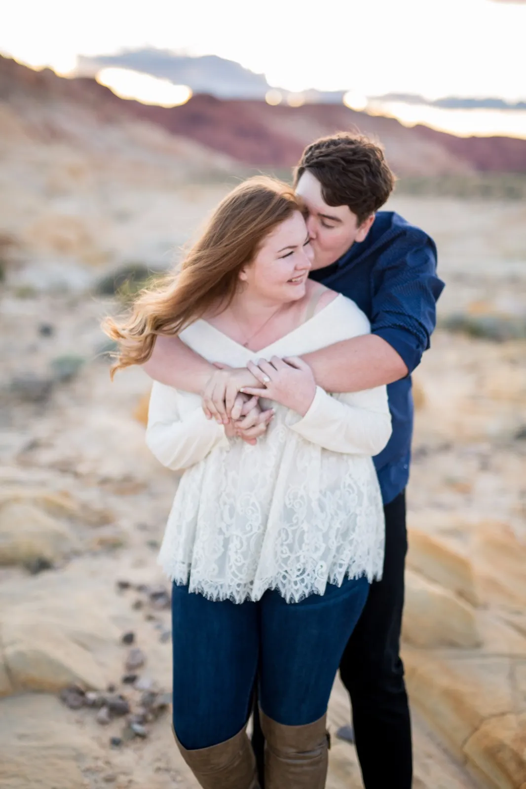 Man hugging his fiancee from behind on pale sandstone while the wind lifts her hair, red hills at dusk