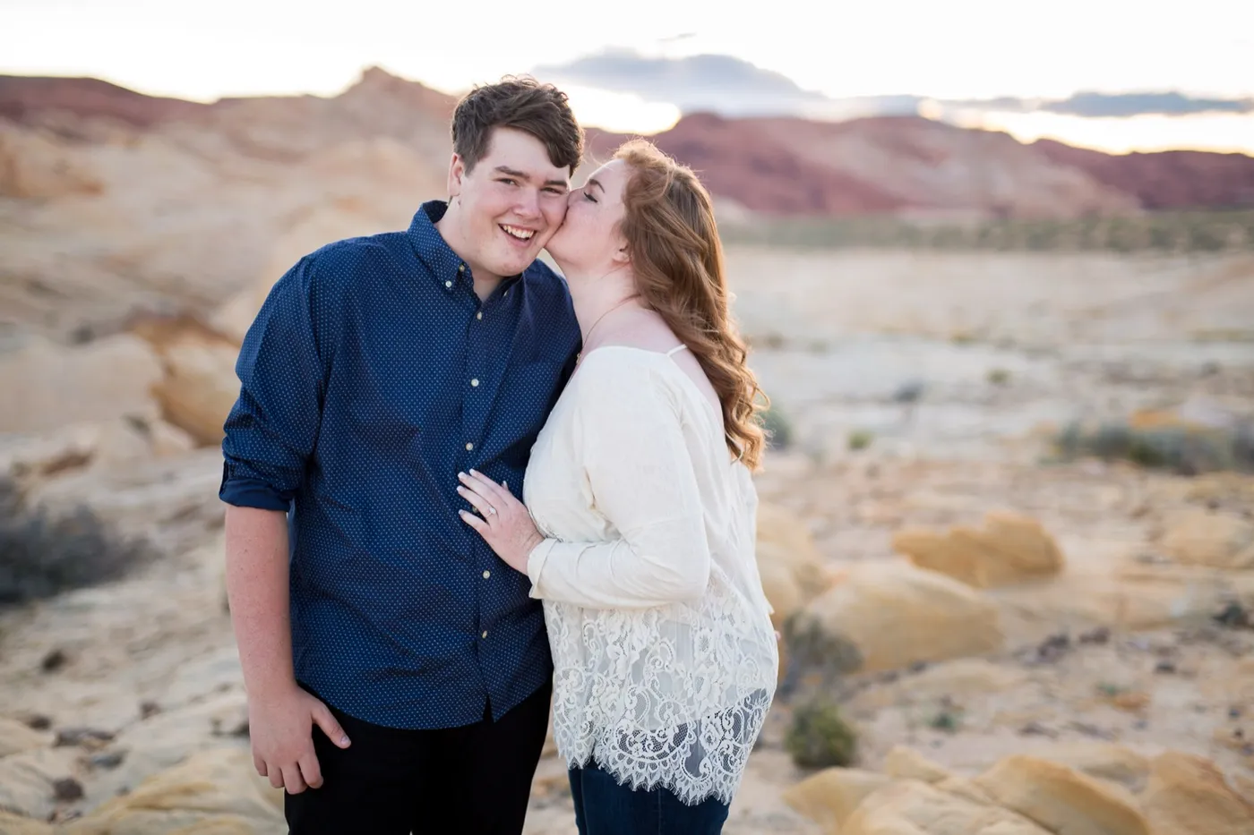 Newly engaged couple on pale sandstone at dusk, she kisses his cheek while he grins at the camera, red hills behind